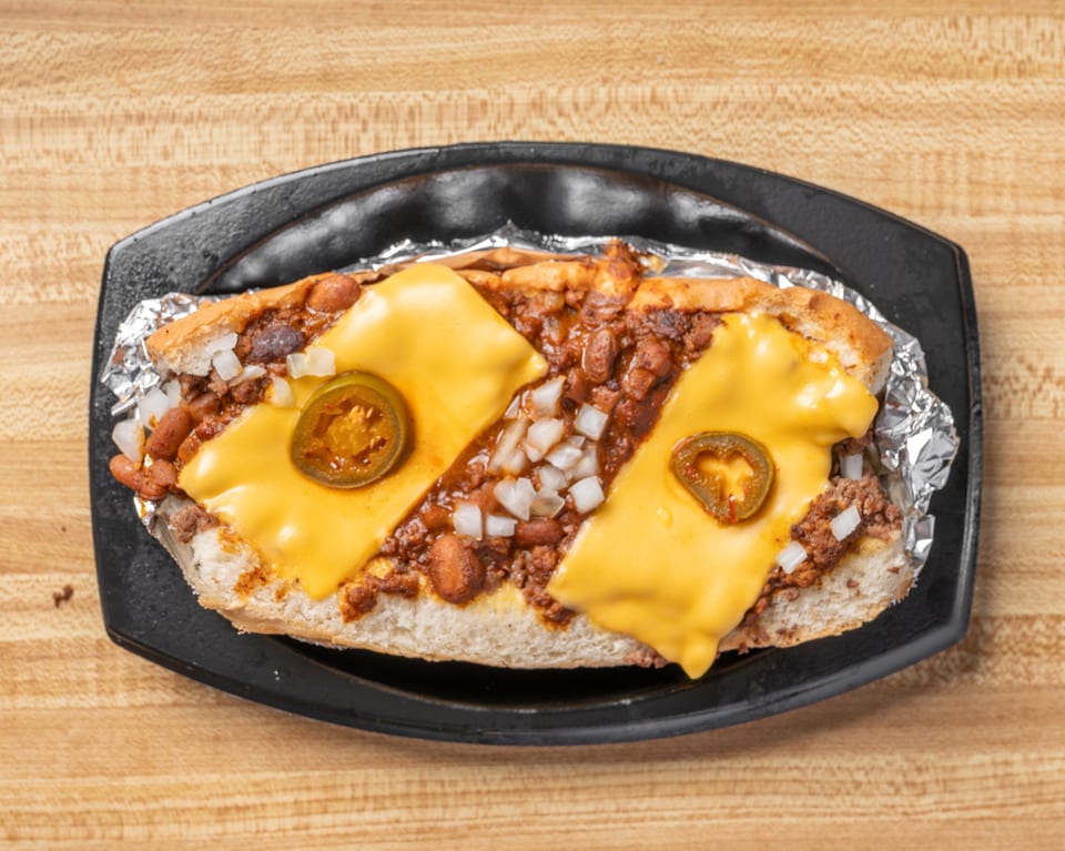 Chili dog with melted cheese, diced onions, and jalapeño slices on a black plate.